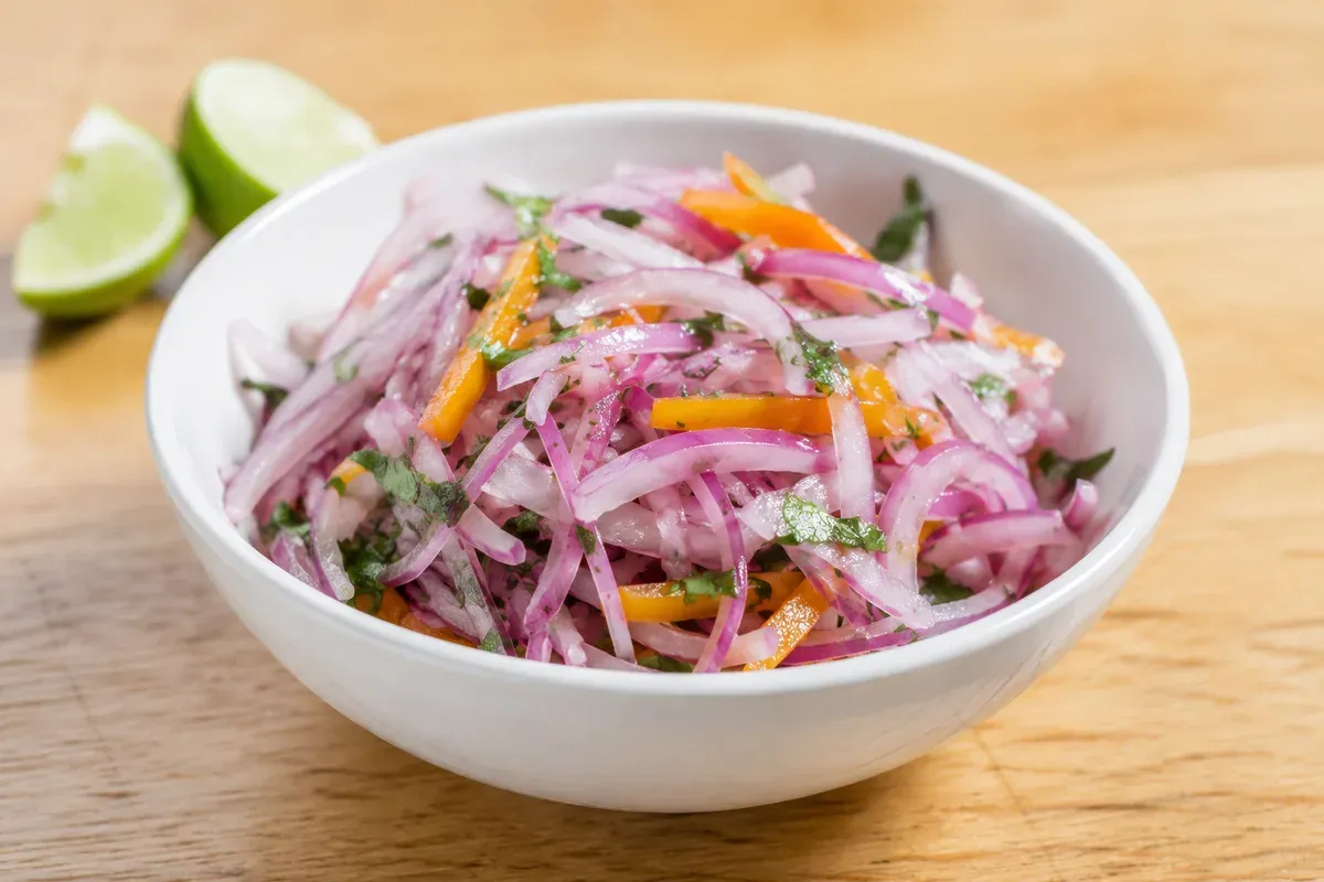 A white bowl of salsa criolla: fine red onion julienne with cilantro and ají strips, glossy with lime, lime halves beside it